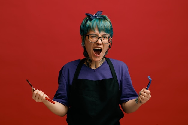 Angry young female barber wearing uniform glasses and headband holding straight razor and shaving razor looking at camera shouting out loudly isolated on red background