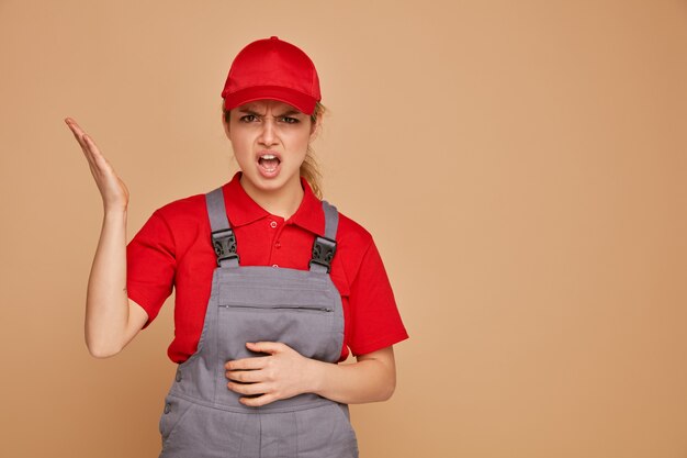 Angry young female construction worker wearing uniform and cap keeping hand on belly showing empty hand 