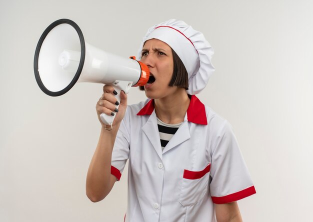 Angry young female cook in chef uniform shouting in loud speaker looking at side isolated on white 