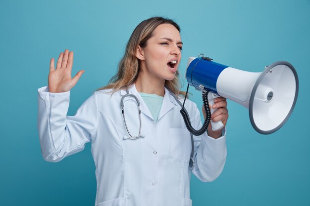 Angry young female doctor wearing medical robe and stethoscope around neck talking by speaker looking at side showing empty hand 