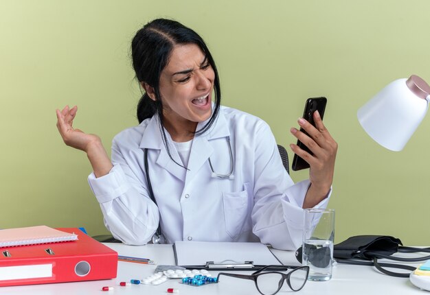 Angry young female doctor wearing medical robe with stethoscope sits at desk with medical tools holding and looking at phone isolated on olive green wall