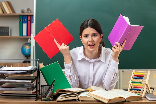 Angry young female teacher holding book sitting at table with school tools in classroom