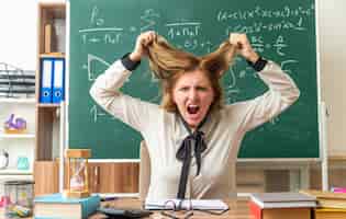 Free photo angry young female teacher sits at table with school supplies grabbed hair in classroom
