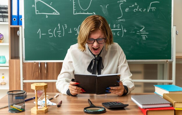 Angry young female teacher sits at table with school tools holding and looking at notebook in classroom