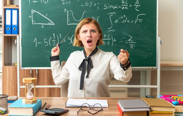 Angry young female teacher sits at table with school tools points at blackboard showing you gesture in classroom