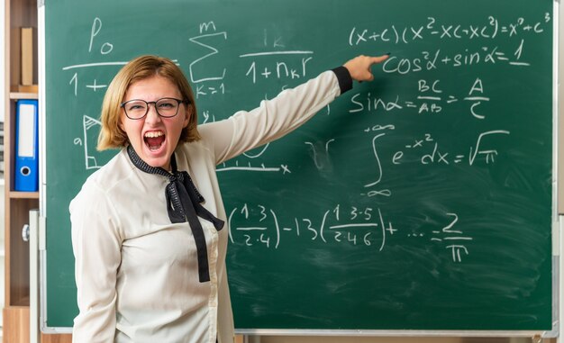 Angry young female teacher wearing glasses standing in front blackboard points at blackboard in classroom