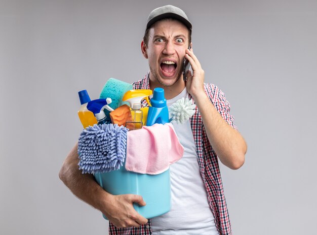 angry young guy cleaner wearing cap holding bucket of cleaning tools speaks on phone isolated on white wall