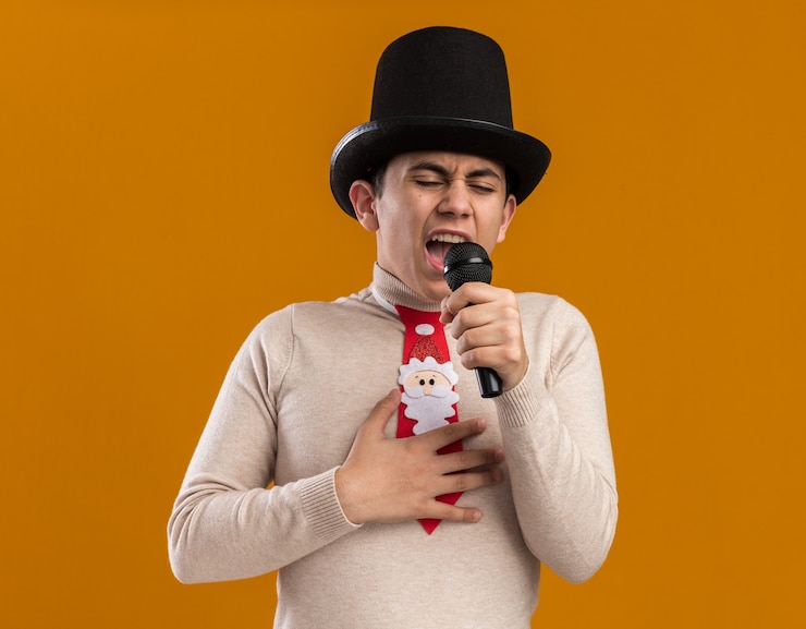 Free Photo angry young guy wearing hat with christmas tie speaks on microphone isolated on yellow wall