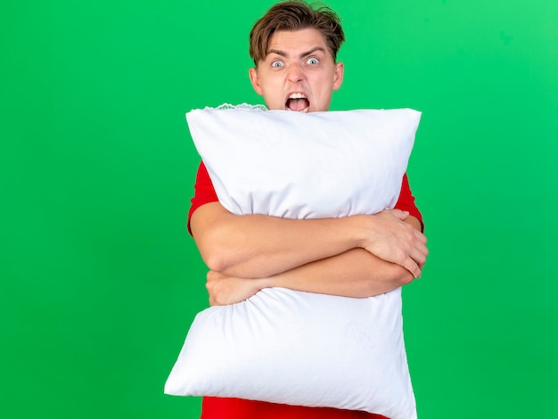 Angry young handsome blonde ill man holding pillow looking at camera from behind it screaming isolated on green background with copy space