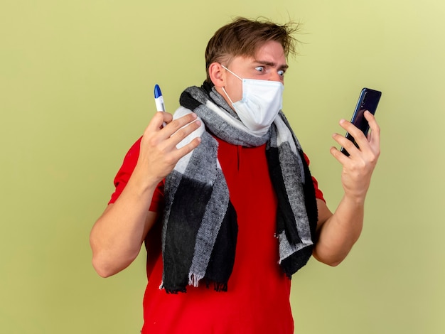 Angry young handsome blonde ill man wearing mask and scarf holding thermometer and mobile phone looking at phone isolated on olive green wall