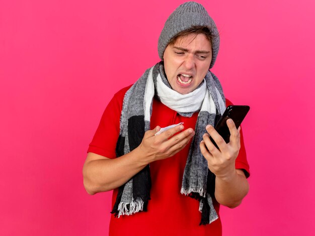 Angry young handsome blonde ill man wearing winter hat and scarf holding mobile phone and napkin looking at phone isolated on pink wall with copy space