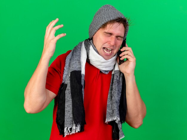 Free photo angry young handsome blonde ill man wearing winter hat and scarf talking on phone keeping hand in air with closed eyes isolated on green wall with copy space