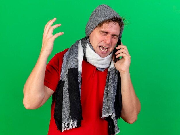 Angry young handsome blonde ill man wearing winter hat and scarf talking on phone keeping hand in air with closed eyes isolated on green wall with copy space