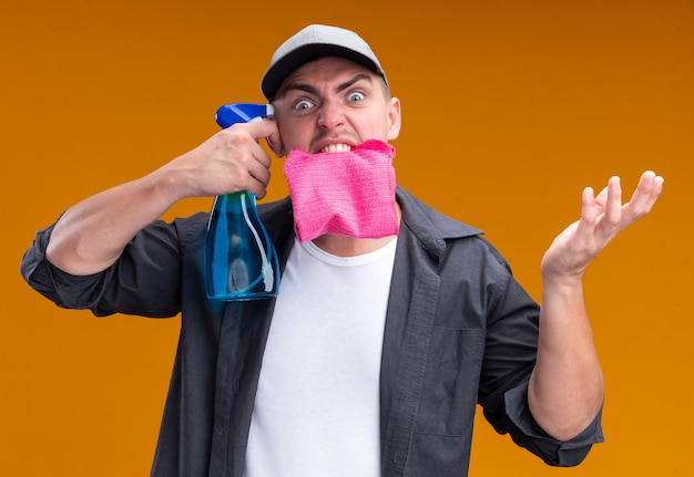 Angry young handsome cleaning guy wearing t-shirt and cap holding rag in mouth putting spray bottle on temple isolated on orange wall