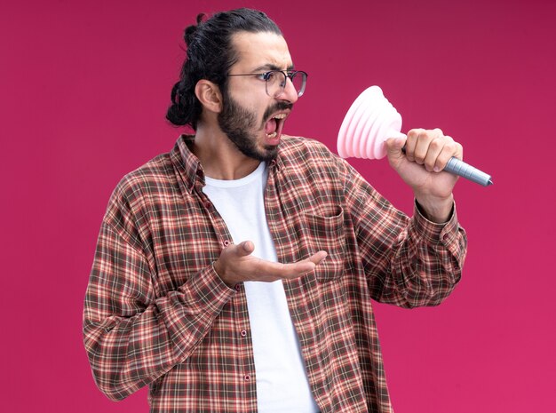 Angry young handsome cleaning guy wearing t-shirt holding and looking at plunger isolated on pink wall