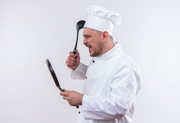 Angry young handsome cook in chef uniform holding frying pan and ladle looking at pan on isolated white wall