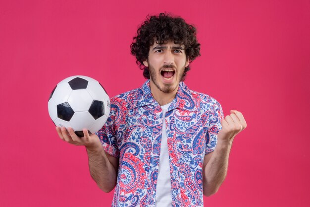 Angry young handsome curly man holding soccer ball with clenched fist on isolated pink space