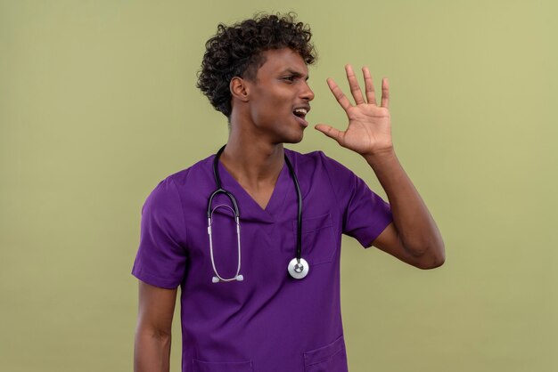 An angry young handsome dark-skinned doctor with curly hair wearing violet uniform with stethoscope calling someone while looking side on a green space