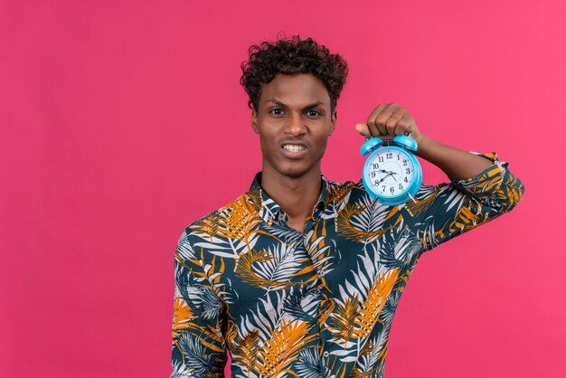 Angry young handsome dark-skinned man with curly hair in leaves printed shirt holding blue alarm clock and showing time 