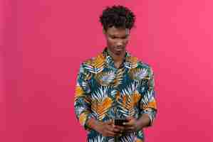 Free photo angry young handsome dark-skinned man with curly hair in leaves printed shirt looking at mobile phone with with a dissenting serious stern expression