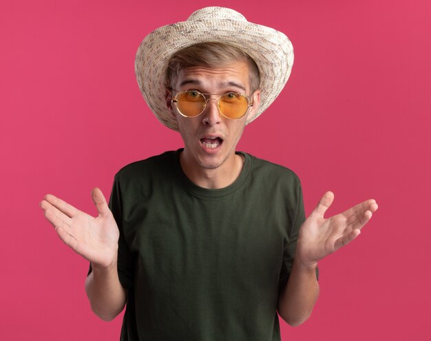 Foto gratuita arrabbiato giovane bel ragazzo indossa camicia verde e occhiali con cappello diffondendo le mani isolate sul muro rosa