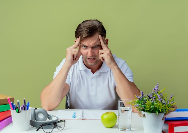 Foto gratuita allievo maschio bello giovane arrabbiato che si siede allo scrittorio con gli strumenti della scuola che mette le mani sulla fronte isolata su verde oliva