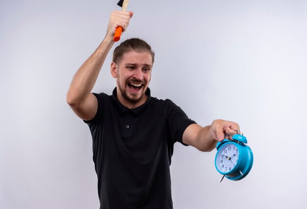 Angry young handsome man in black polo shirt with a hammer going to break alarm clock in his hand with aggressive expression on face standing over white background