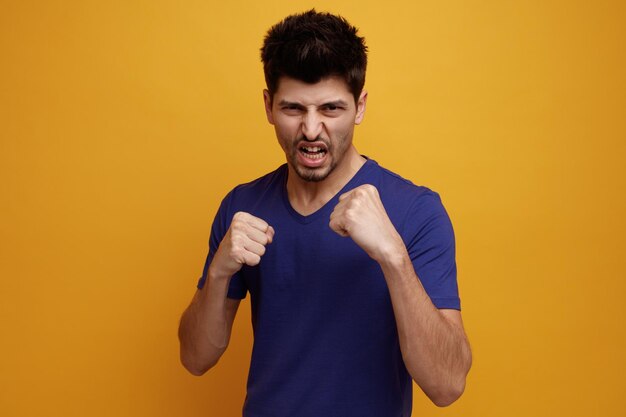 Angry young handsome man looking at camera doing boxing gesture on yellow background