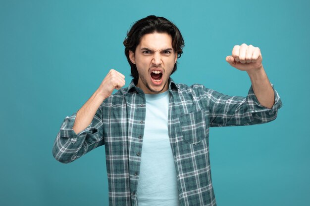 angry young handsome man looking at camera keeping fist in air showing punch gesture isolated on blue background