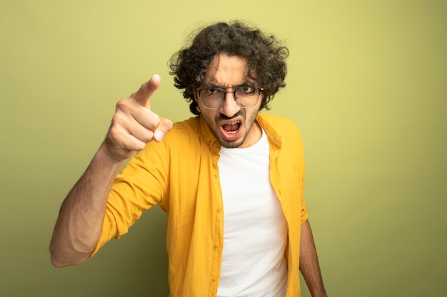 Angry young handsome man wearing glasses looking and pointing at front isolated on olive green wall