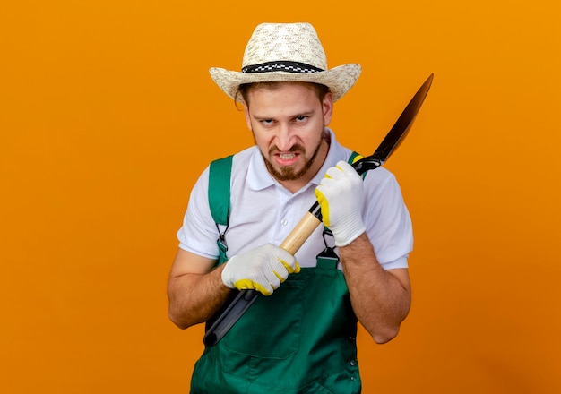 Сердитый молодой красивый славянский садовник в униформе в шляпе и садовых перчатках, держащий лопату, выглядит изолированно