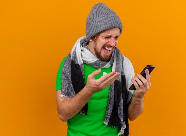 冬の帽子とスカーフを身に着けて、コピースペースのあるオレンジ色の壁に隔離されたナプキンを手に携帯電話を指している携帯電話を見て怒っている若いハンサムなスラブ病の男
