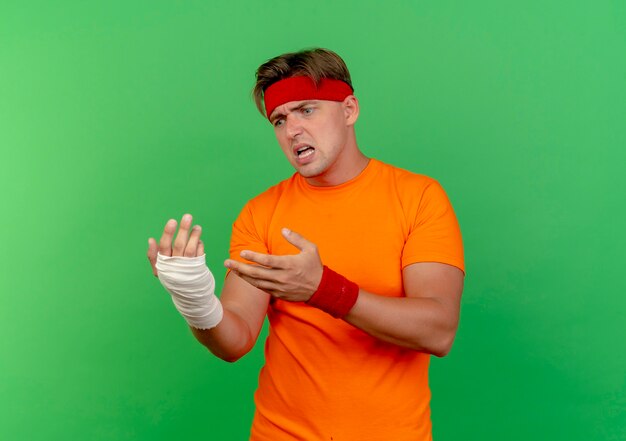 Angry young handsome sporty man wearing headband and wristbands looking and pointing at his injured wrist wrapped with bandage isolated on green  with copy space