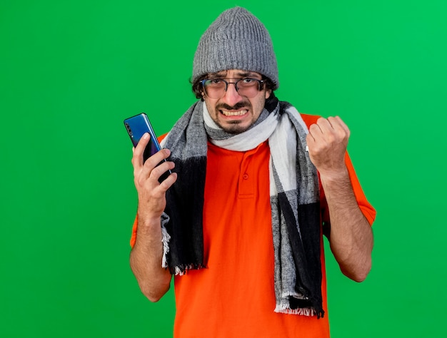 Сердитый молодой больной человек в очках, зимней шапке и шарфе держит мобильный телефон и салфетку, глядя вперед, изолированную на зеленой стене