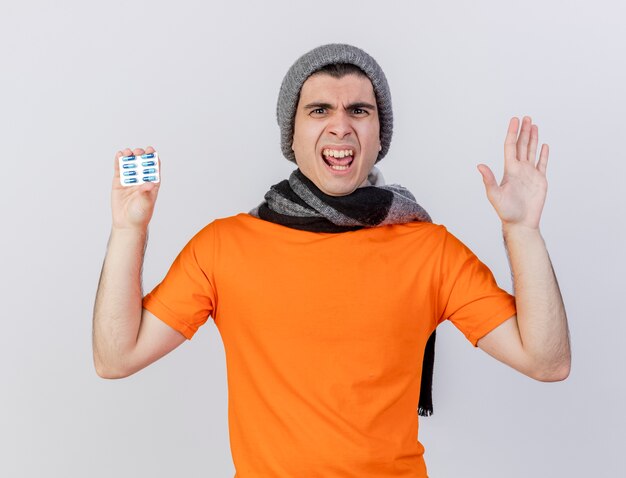 Angry young ill man wearing winter hat with scarf holding pills and showing stop gesture isolated on white