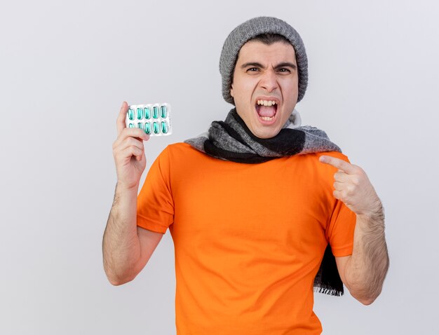 Angry young ill man wearing winter hat with scarf holding and points at pills isolated on white