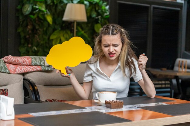 Angry young lady holding yellow idea board and sitting at the restaurant