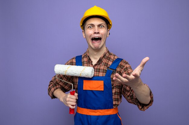 Angry young male builder wearing uniform holding roller brush