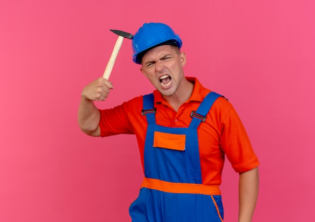 Angry young male builder wearing uniform and safety helmet putting hammer on helmet 