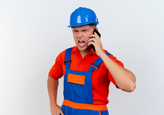 Angry young male builder wearing uniform and safety helmet speaks on phone 