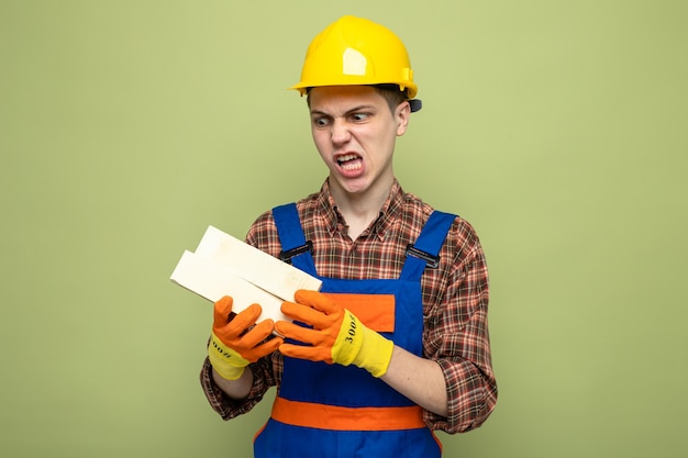 Angry young male builder wearing uniform with gloves holding and looking at bricks 