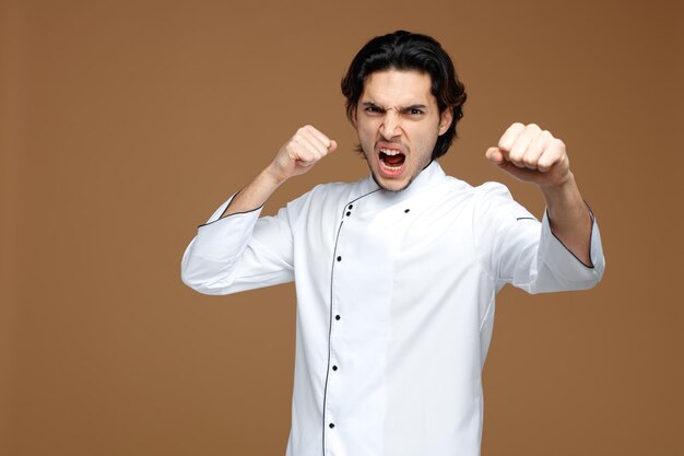 angry young male chef wearing uniform looking at camera stretching fist out towards camera showing punch gesture isolated on brown background