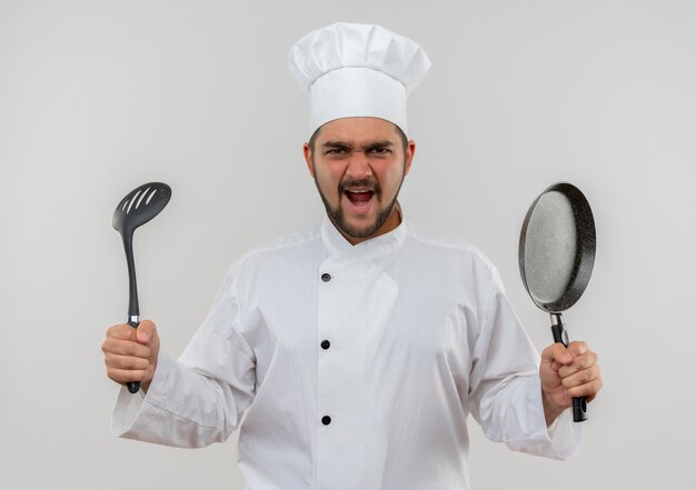 Angry young male cook in chef uniform holding slotted spoon and frying pan isolated on white wall