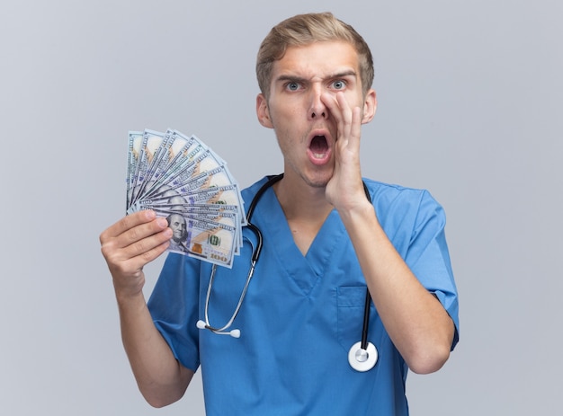 Angry young male doctor wearing doctor uniform with stethoscope holding cash calling someone isolated on white wall