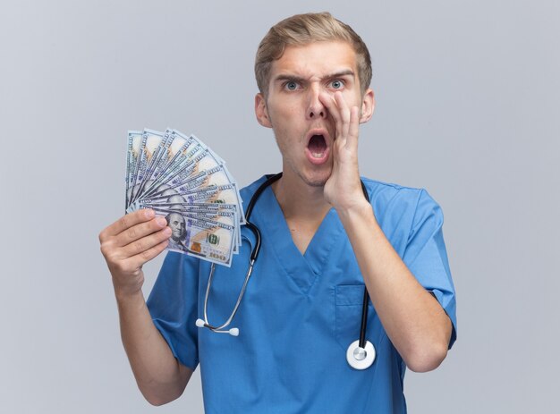 Angry young male doctor wearing doctor uniform with stethoscope holding cash calling someone isolated on white wall