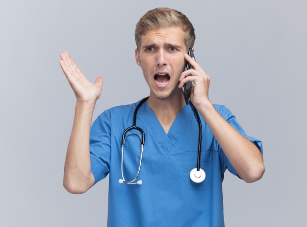 Сердитый молодой мужчина-врач в униформе врача со стетоскопом говорит по телефону, протягивая руку, изолированную на белой стене