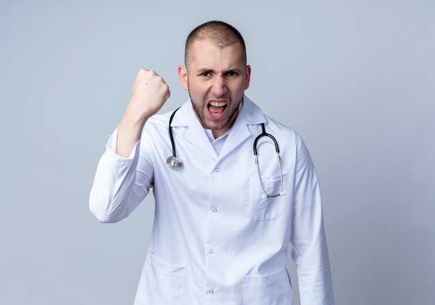 Angry young male doctor wearing medical robe and stethoscope around his neck raising fist isolated on white  with copy space