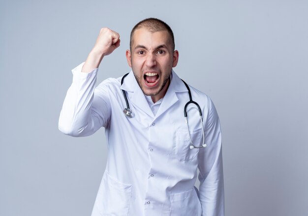 Angry young male doctor wearing medical robe and stethoscope around his neck raising his fist and screaming isolated on white  with copy space