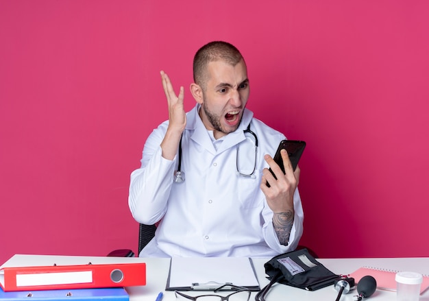 Arrabbiato giovane medico maschio indossa veste medica e stetoscopio seduto alla scrivania con strumenti di lavoro tenendo e guardando il telefono cellulare con la mano alzata isolata sul rosa