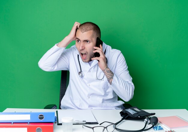 Angry young male doctor wearing medical robe and stethoscope sitting at desk with work tools talking on phone putting hand on head looking at side isolated on green 